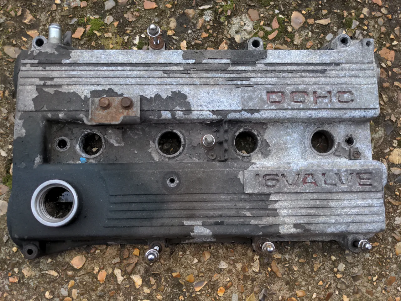 A Mazda B6T rocker cover, clearly very old. About half of it is covered in a mix of flaky black paint and oil, and it is not clear where the border between the two is. The parts that are not oily or greasy have surface corrosion.
