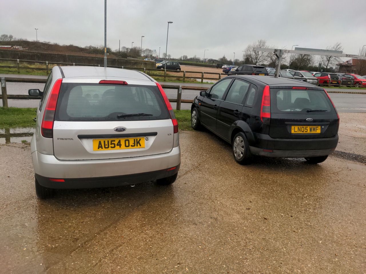 Two fifth-generation Ford Fiestas parked next to each other in a car park; mine is silver, the other is black, neither is very clean and it is a drab overcast day.