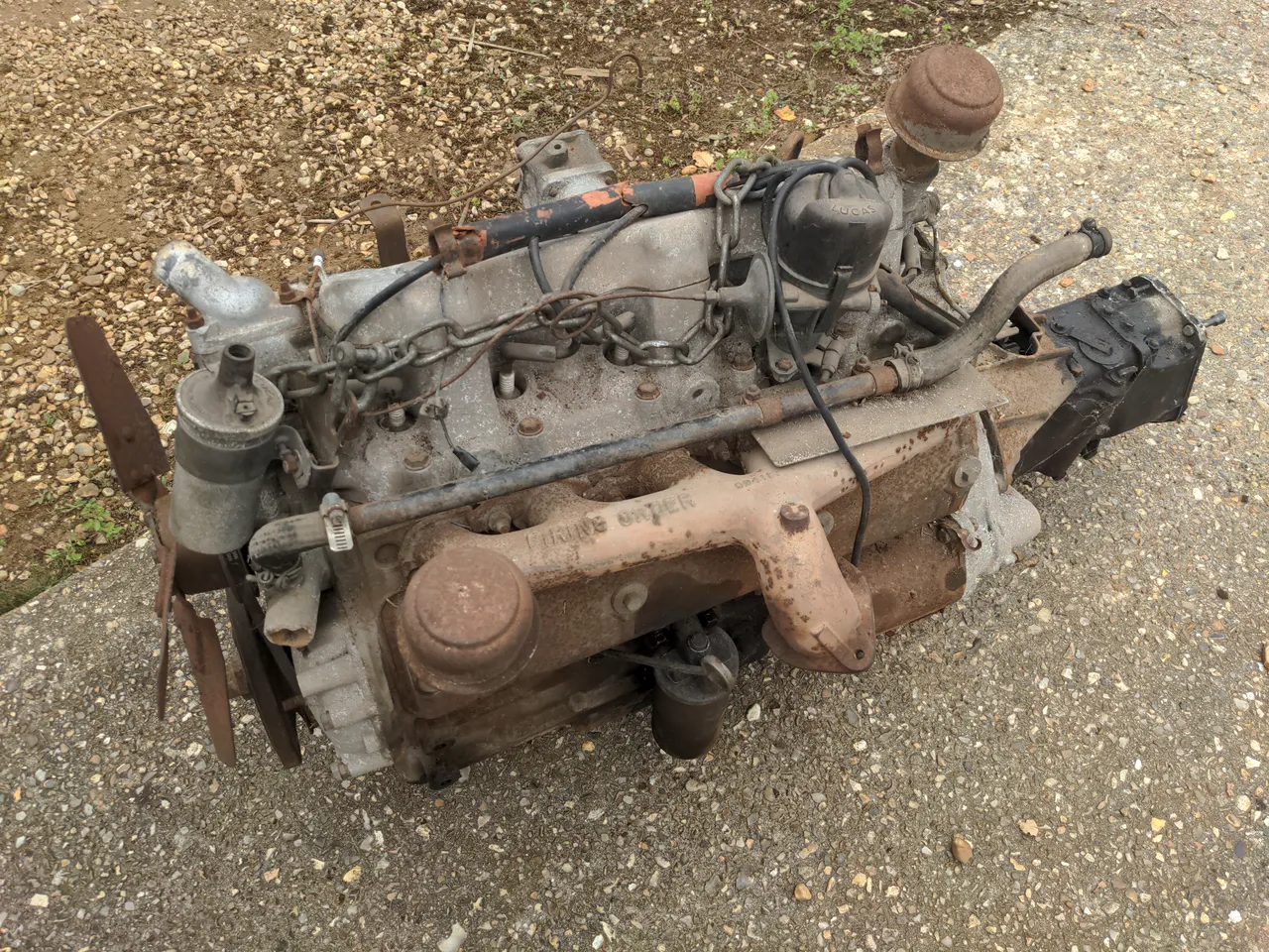 A slightly-rusty Rover P5 engine sitting on a concrete floor. It is a large six-cylinder engine of an ancient design. It has the transmission attached.