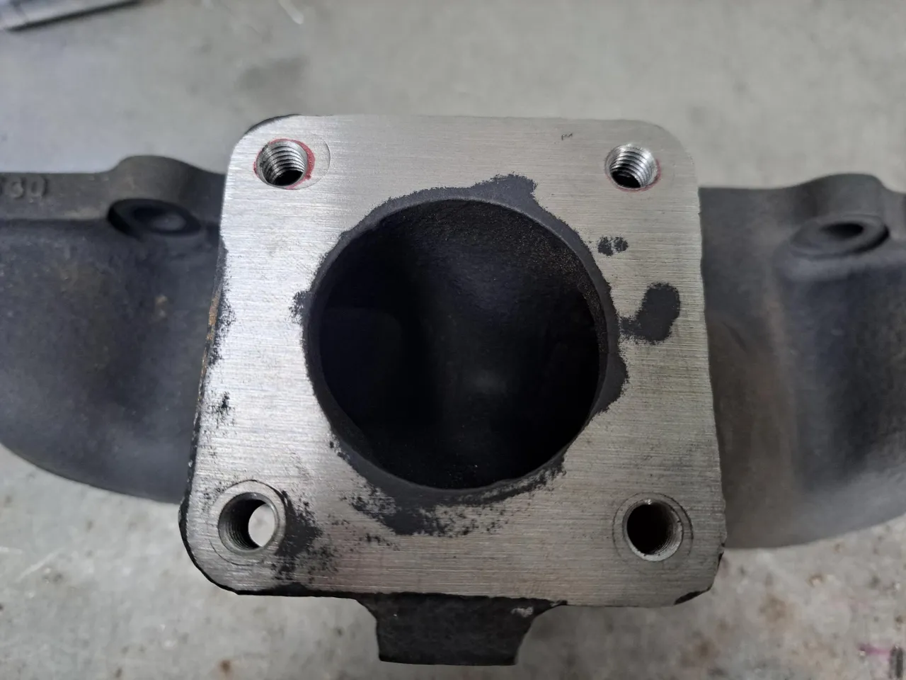 The turbo end of an exhaust manifold. It has been ground flush. The parts where it has been drilled out are visible via a tiny black outline, and these have had plugs rammed into them. Four M8-threaded holes have been drilled into it very precisely.