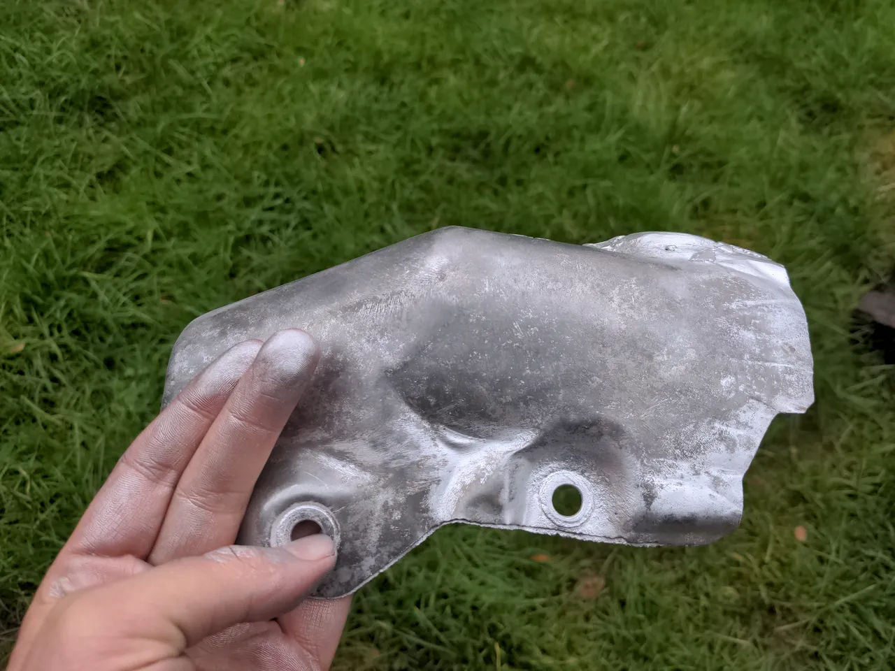 The heat shield in my hands. Two fingers are visible, coated in silver. The paint has almost entirely come off the heat shield.