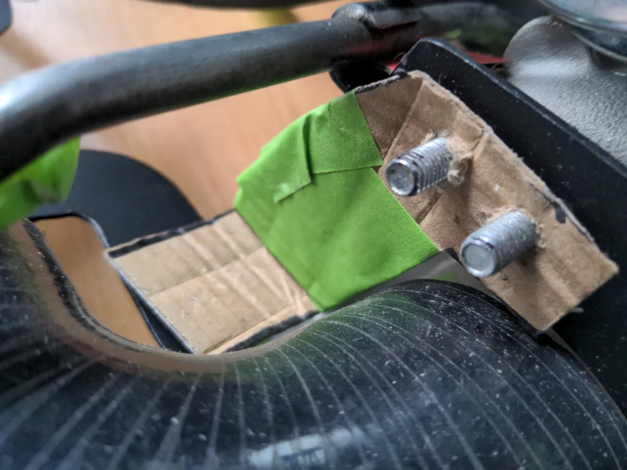 A crude prototype of a bracket, cut out of cardboard. Some green masking tape reinforces the cardboard. On one end, the two M6 bolts from earlier poke through the cardboard.