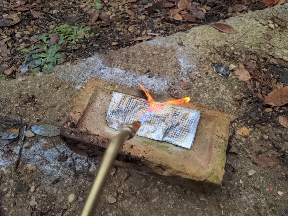 An oxypropane torch directed at some reflective material which is glued to a piece of steel. This is sitting on a brick.