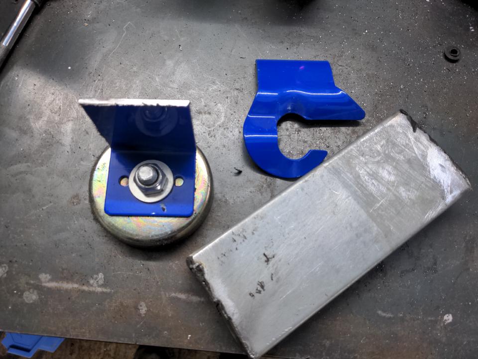 A section of galvanised steel C-section, cut in such a way that the ends have been folded over to form a box with one side missing. A magnetic welding clamp sits on the same workbench, with half of it cut off, leaving just the big magnet (to attach it to a metal surface) with a right-angled piece of steel bolted to it.