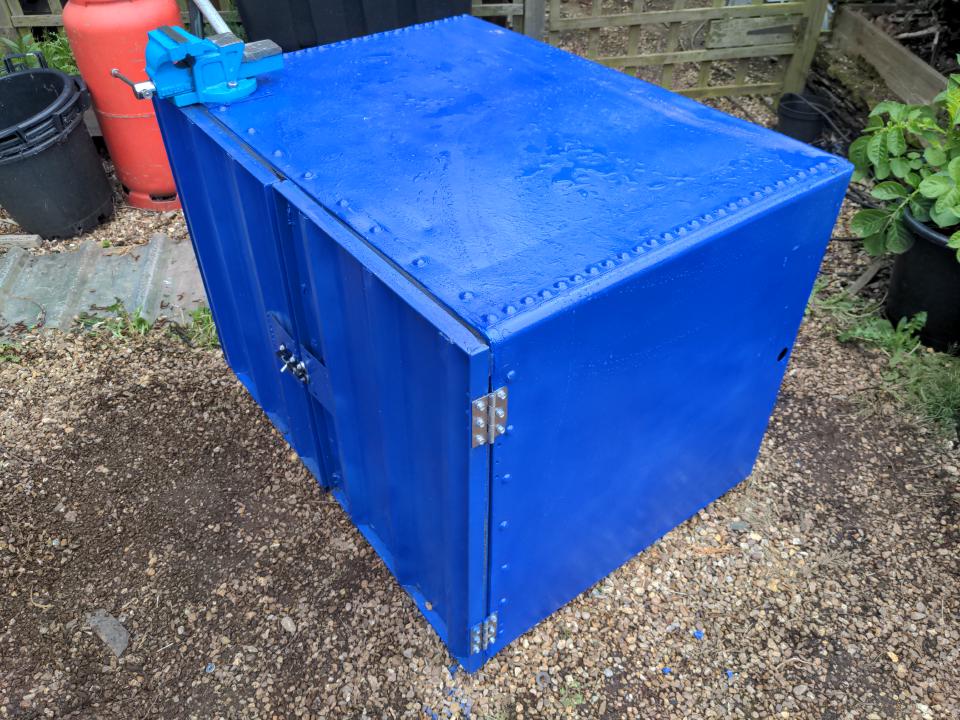 My outdoor workbench/cabinet. It is painted a vivid medium blue called Jenolite Ultramarine Blue Gloss. This used to be a water tank as fitted into the loft of a typical house in the early to mid 20th century; it has doors on the front where it used to be an open top. A blue vice is attached to it.