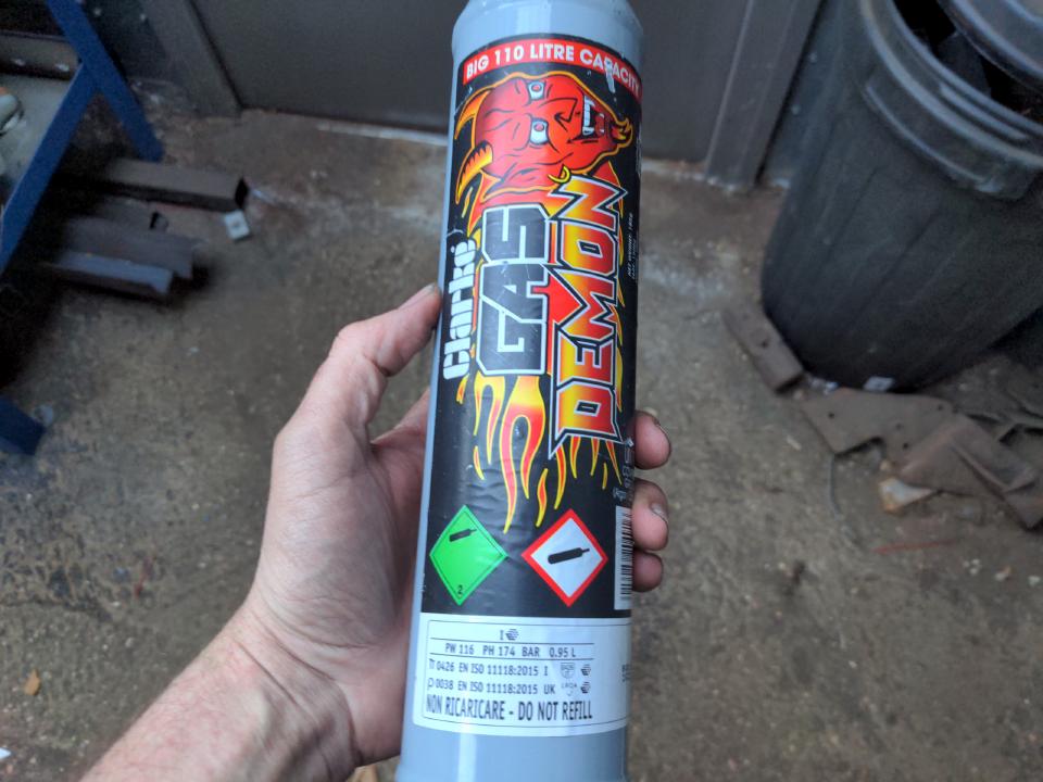 A disposable bottle of argon/CO2 shield gas; it is in my hand. It is about a foot and a bit long and its diameter fits in my hand. It has GAS DEMON and various flame graphics on it.