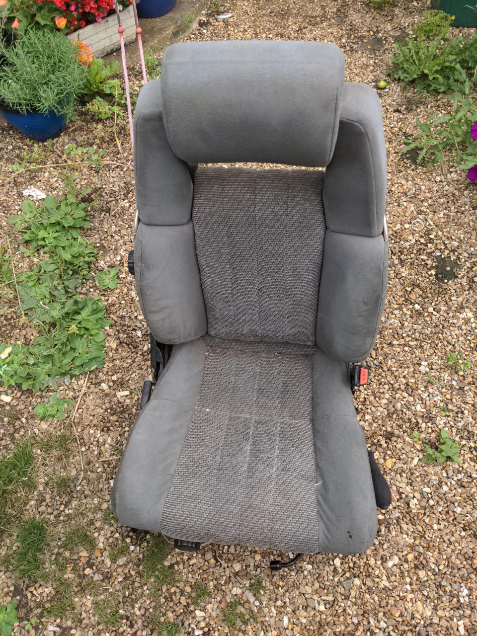 A front seat from a 1985 Toyota Supra. It is almost a bucket-like seat, with very generous side supports. It is clad in a very textured grey-black-mottled fabric in the middle and smooth plain grey fabric on the bolsters and headrests.