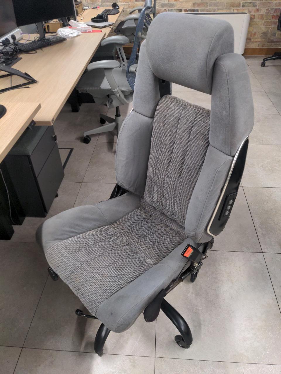 A grey cloth mid-80s Toyota Supra front seat with a swivel chair base at an office, with Aeron chairs visible in the background.