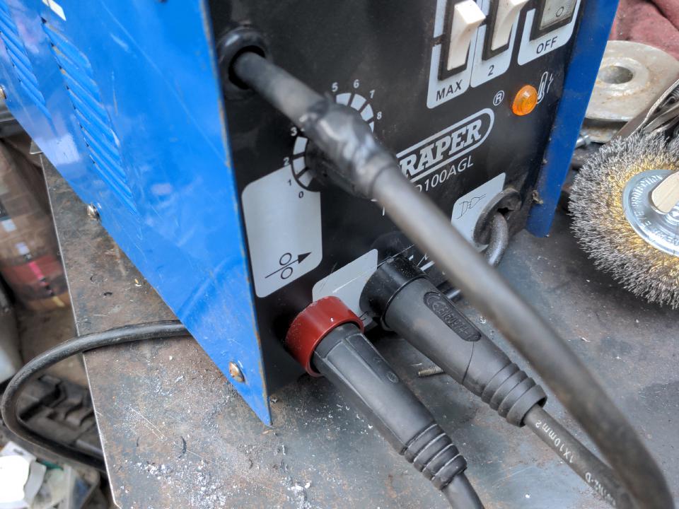 A front view of my modified welder. A cable comes out the front near the top through a grommet, and immediately goes to a black (positive) Dinse-type plug; this is connected directly to the torch. This cable shows evidence of being cut-and-shut; a bulge with heat shrink is visible. As before, the return is connected to a red (positive) Dinse socket.