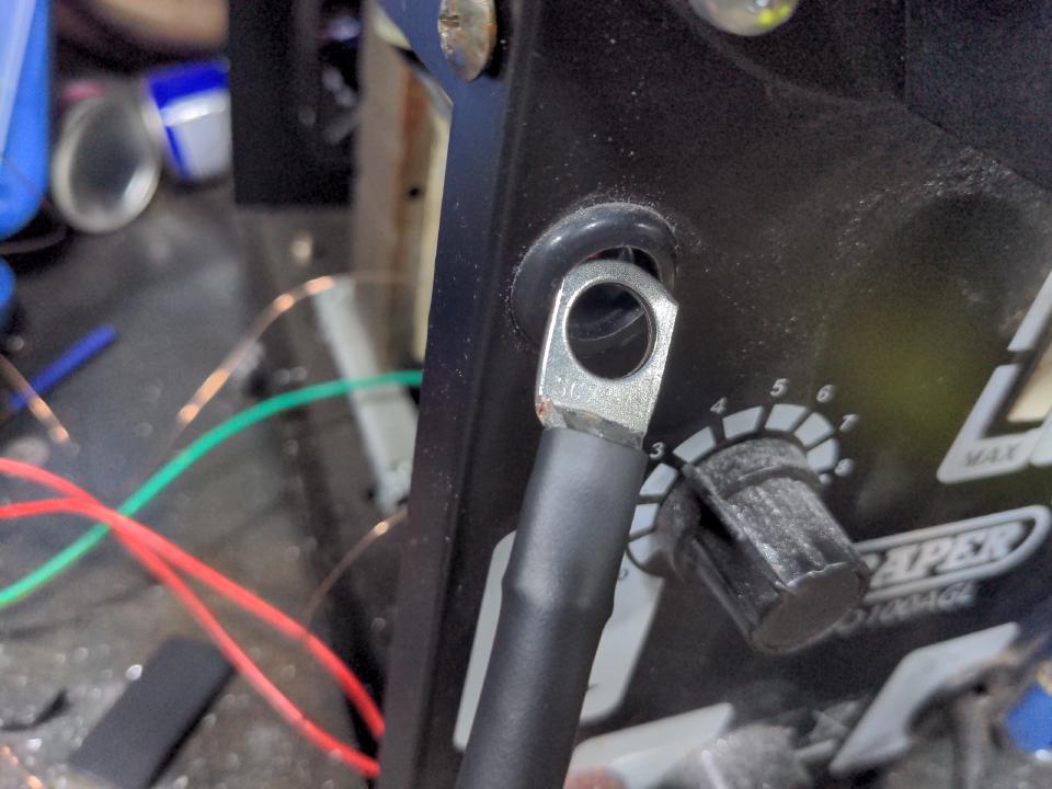 A cable with a ring terminal attached, sitting next to a hole that the cable is meant to feed through. The problem displayed is that a) the connector is meant to be on the OTHER SIDE of the hole and b) the connector is clearly too large to fit through the hole. What I should have done is fed the cable through the hole THEN fitted the connector, but I did not.