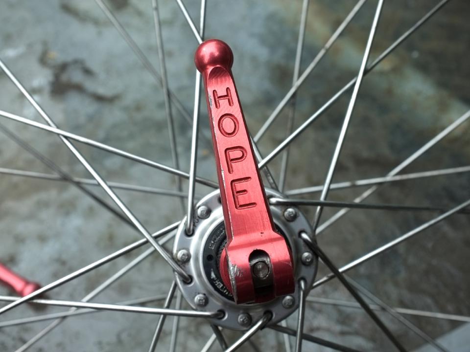 A Hope titanium QR skewer. It is red and has H-O-P-E written down its length vertically. The shape slightly tapers towards the end and ends with a small ball shape.