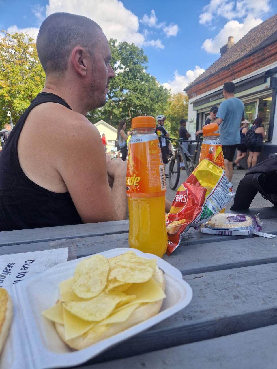 A photograph of me at an outdoor cafe popular with cyclists at High Beach. Lucozade and crisps are on the table while I watch the scene.