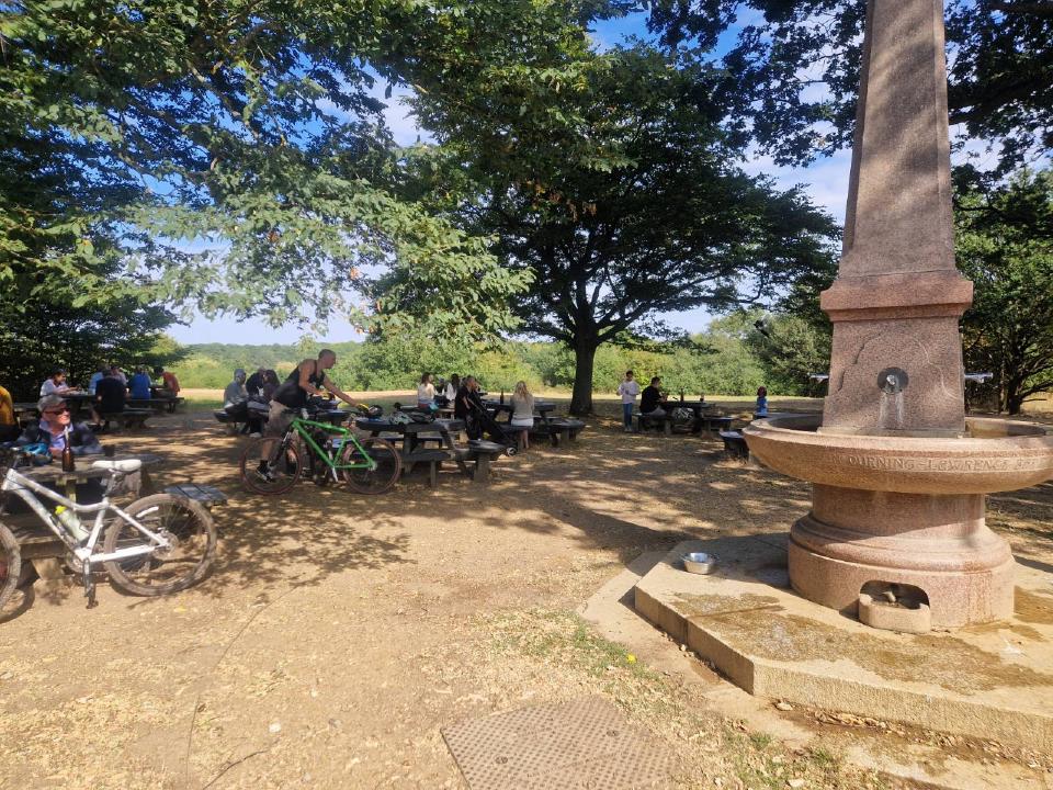 Taking a pause in a nicely shaded outdoor area under some trees, with a water fountain in the foreground. I am visible in a small pack of people at the tables.
