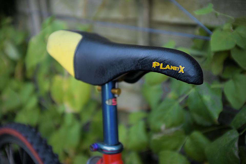 A Planet X downhill-style seat; it is black with yellow kevlar corners and is somewhat extended downwards where your thighs are.