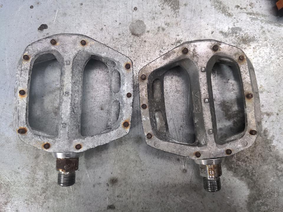 Two DMR V12 pedals. They are aluminium pedals of the flat design similar to BMX bikes of the 1980s and 1990s. The aluminium has corroded to white, and the steel studs (to grip the sole of your shoes) have rusted to orange.