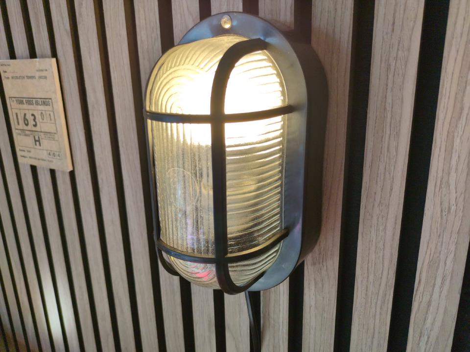 A bulkhead light mounted to a wood slat wall; the cable exits out the bottom and disappears into the gaps between the slats.