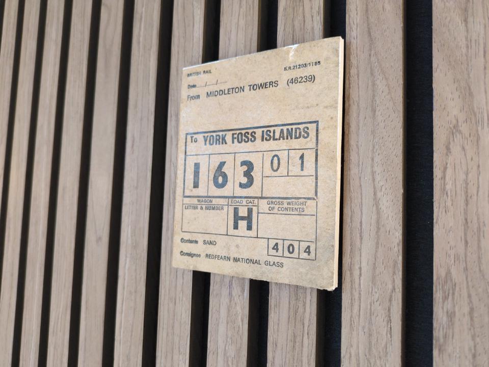 A small paper label, indicating a sand train destination of York Foss Islands, mounted on a foam board on a wood slat wall.